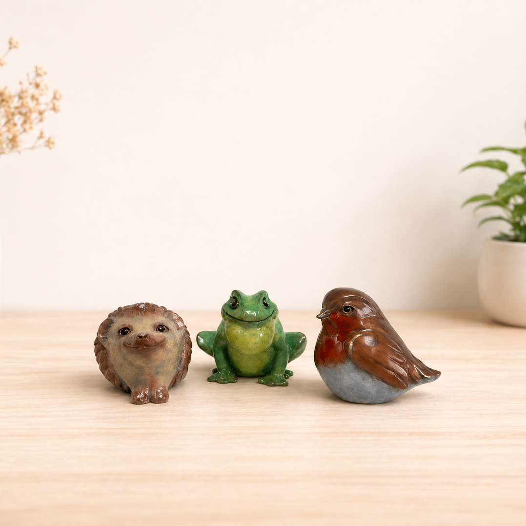 Three small animal figurines (hedgehog, frog, and robin) on a wooden surface with plants in the background.